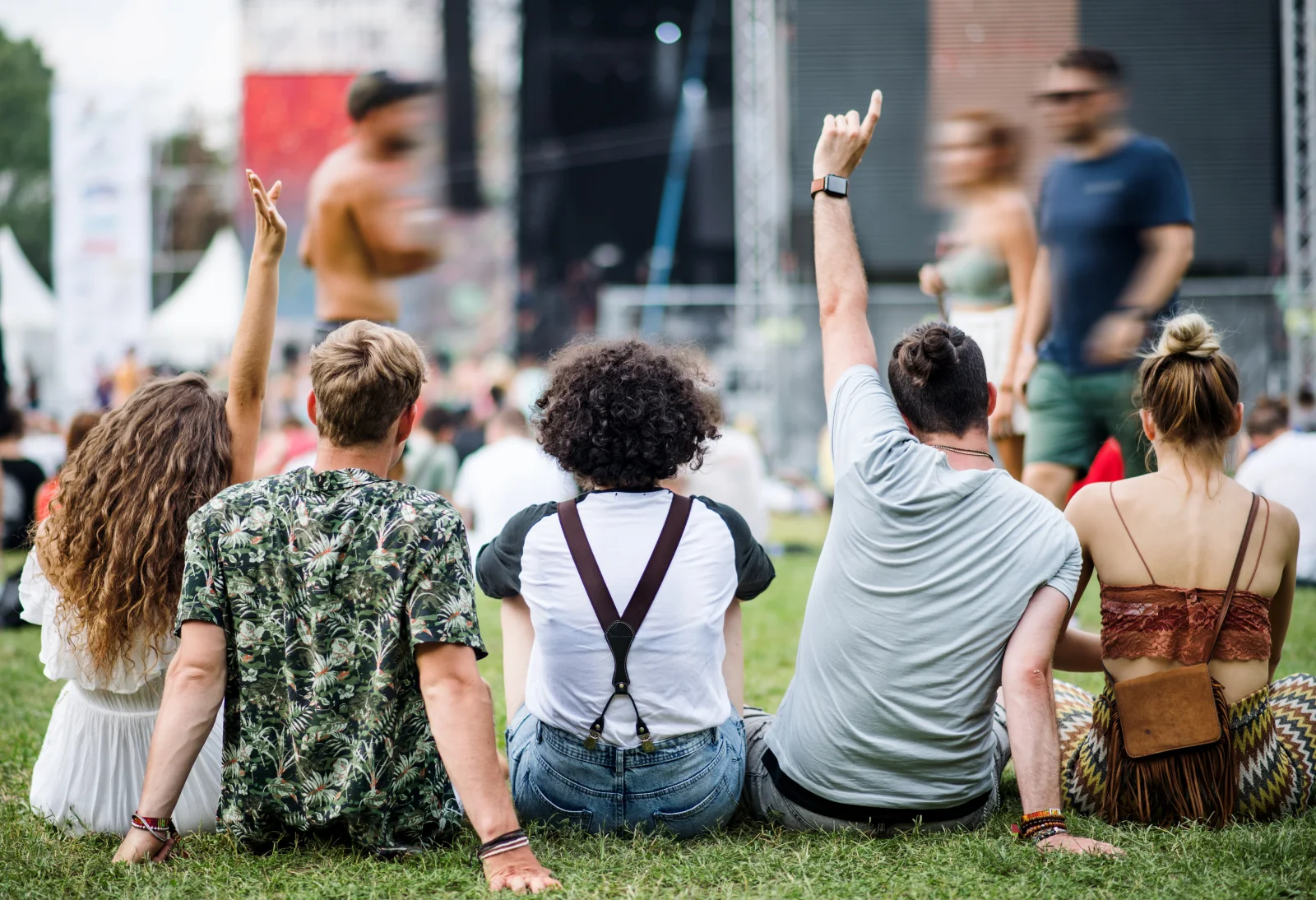 Diverse adult friends viewed from behind, sitting on grass and watching a daytime festival